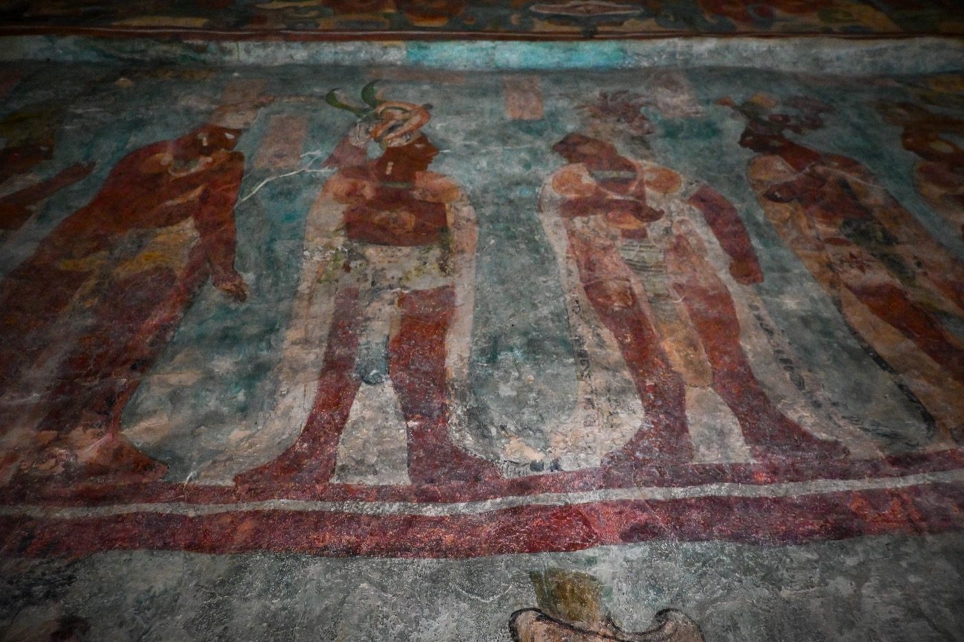 The original, weathered Bonampak murals still on the walls at the archaeological site, showing faded but legible painted figures in procession against a blue-green background.