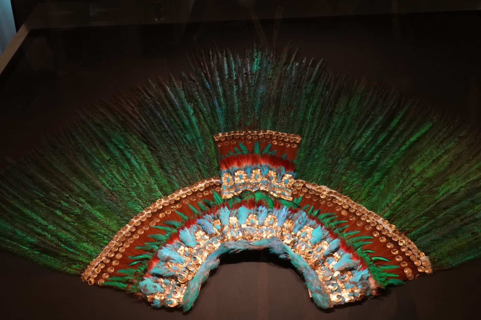 Moctezuma's feather headdress on display at the Weltmuseum in Vienna, showing concentric layers of long green quetzal tail feathers, blue cotinga feathers, and small gold ornaments arranged in a fan shape.