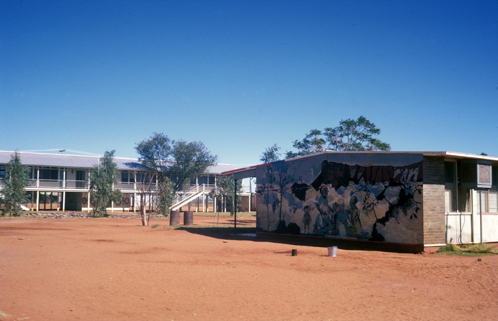 A 1972 photograph of the Papunya settlement school in the Northern Territory, showing low brick and timber buildings around a red dirt yard, with a painted mural visible on the end wall of one building.