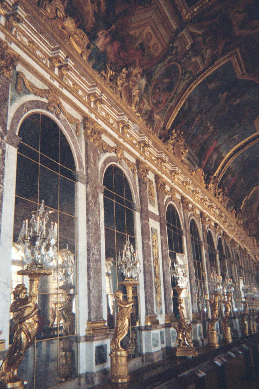A row of tall arched mirrors set between marble pilasters in the Hall of Mirrors at Versailles, with gilded figures holding candelabra in front and a painted vault overhead.